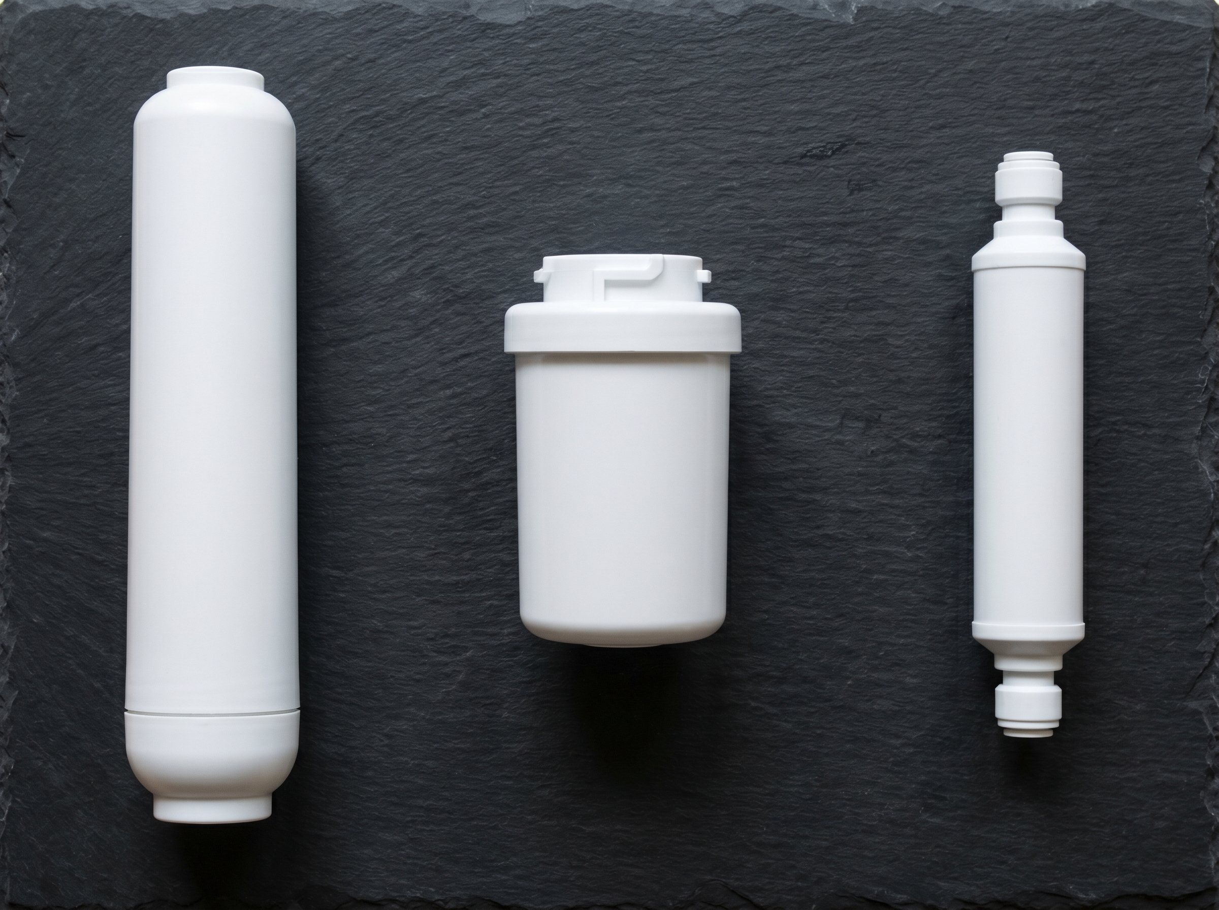 Three fridge water filter cartridges laid side by side on dark slate: an internal twist-in cylinder, a shorter push-fit door cartridge, and a slim inline external filter